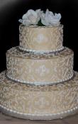 Champagne/gold buttercream iced 3 tiered wedding cake, Contrasting white scroll work on all tiers, with an oval monogram centered and Gardenia toppers. (This cake can serve receptions with 90-220 expected guests)